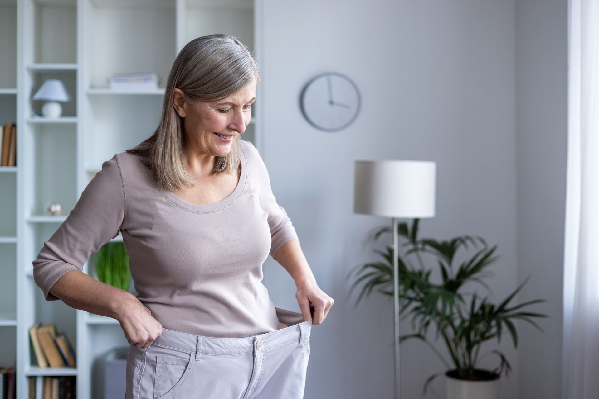 Smiling senior woman feels accomplished as she admires her weight loss progress in her large pants, standing in a cozy room, symbolizing healthy lifestyle and personal achievement.